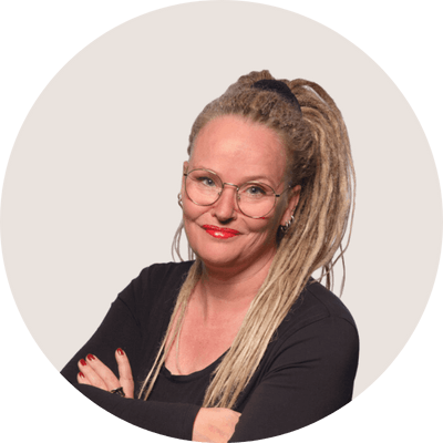 Portrait of Anne-Mieke Bovelett, a woman wearing long blonde dreads in a ponytail, big rounded silver rimmed glasses, a black shirt, with a subtle smirk on her face.